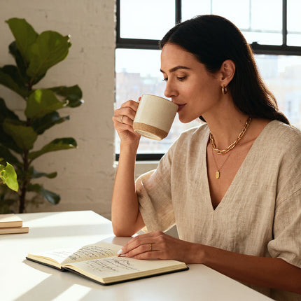 Woman drinking from a mug and reading a book in a bright room with a plant.